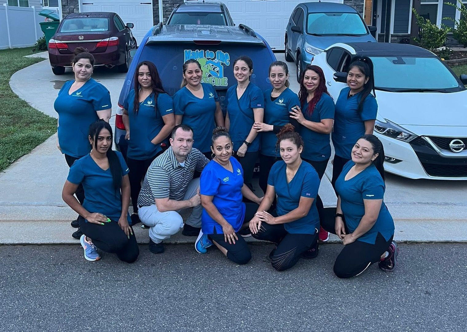 A group of eleven people in teal scrubs posing for a photo on a driveway in front of two cars.