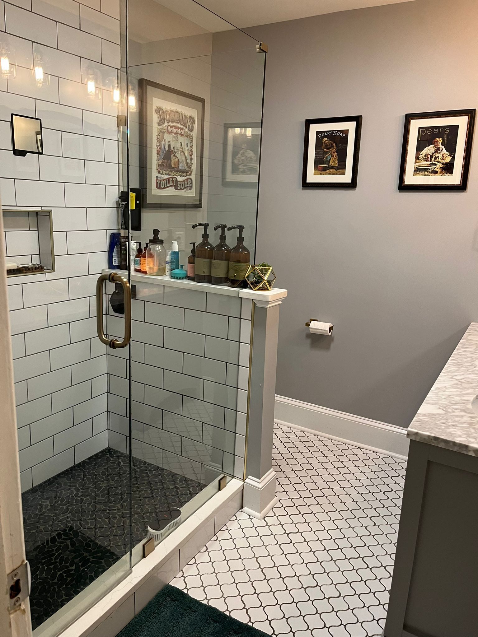A bathroom with white subway tile shower, geometric patterned floor tiles, gray walls, and two framed wall pictures.