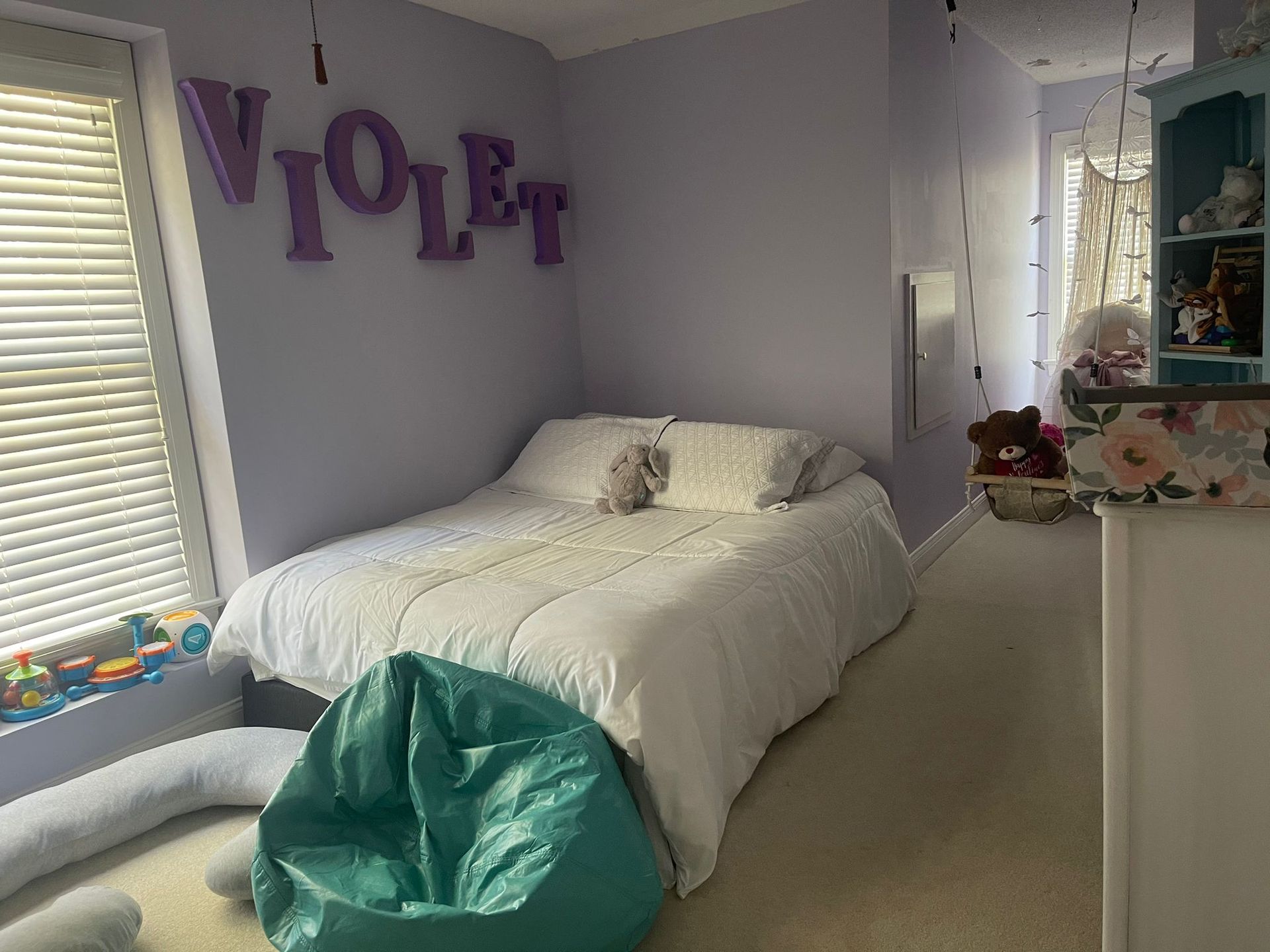 A bedroom with light purple walls, a bed with white bedding, a teal beanbag chair, and the name 