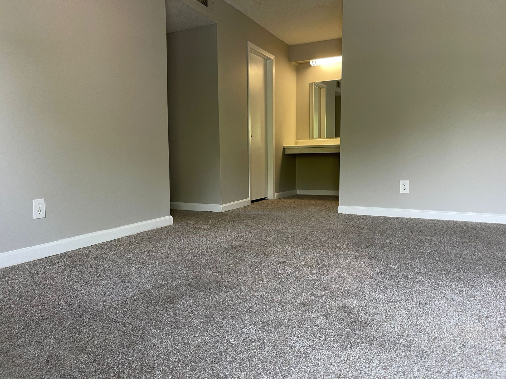 An interior room featuring light gray walls, gray speckled carpeting, a doorway to the left, and a vanity area ahead.