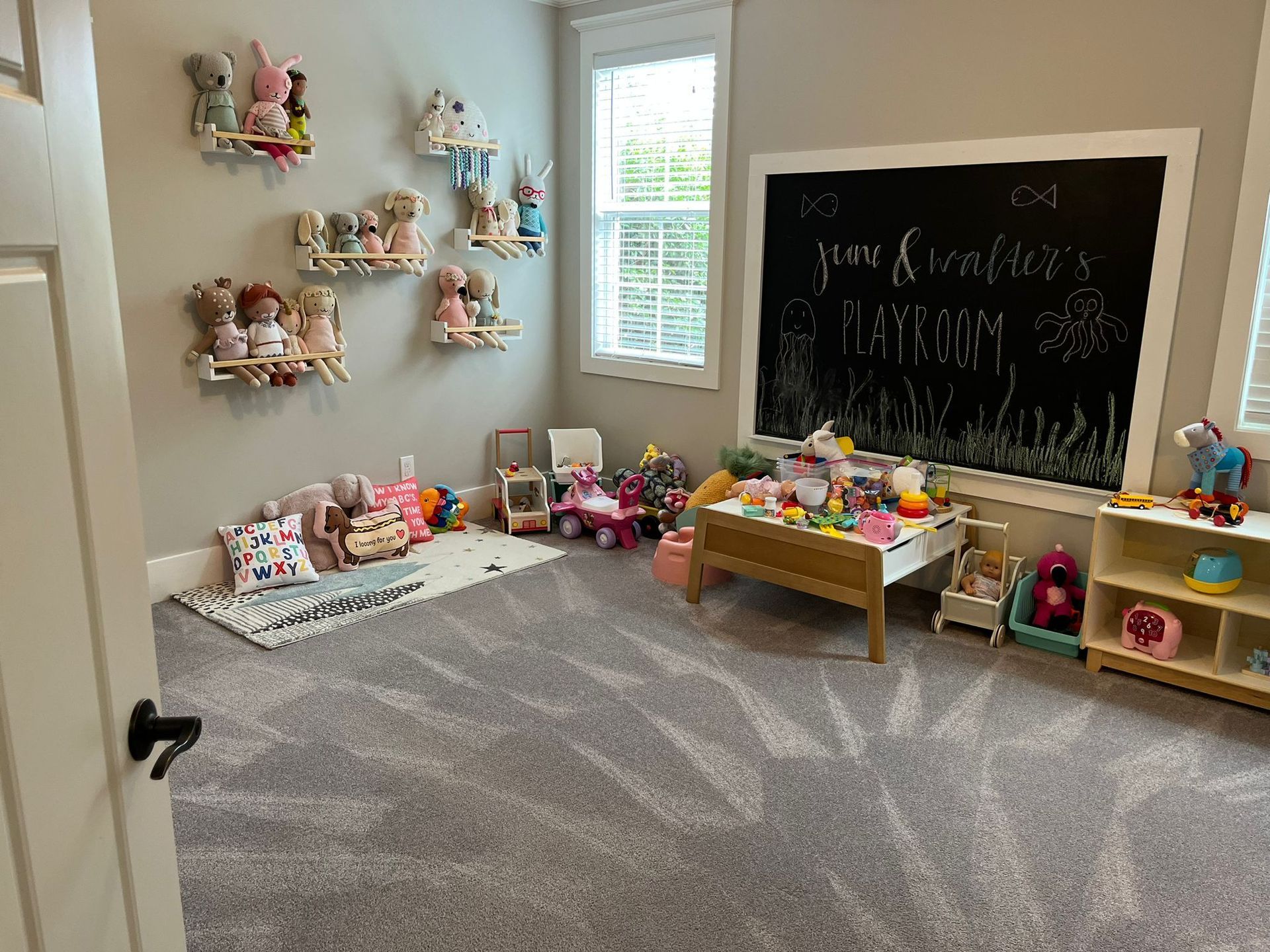 A cozy playroom with gray carpet, walls featuring plush toy displays, a large wall-mounted chalkboard, and toy furniture.