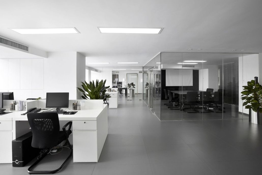A modern office with white desks, black chairs, a glass-walled conference room, and plants on a gray floor.