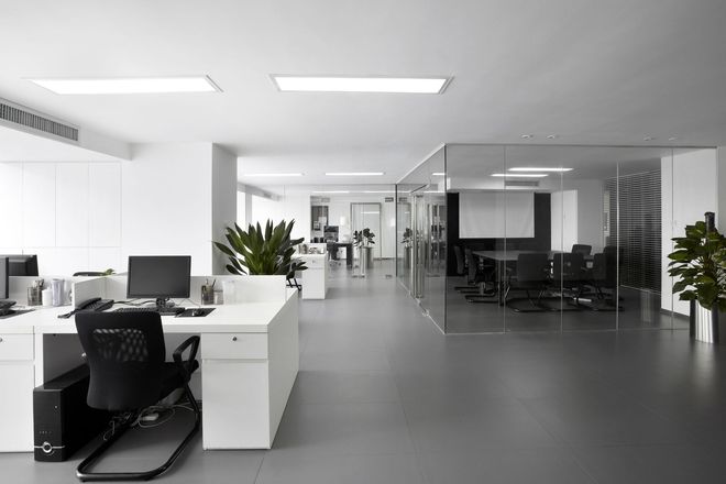 A modern office with white desks, black chairs, a glass-walled conference room, and plants on a gray floor.