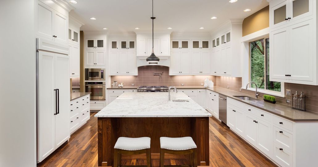 A spacious, modern kitchen with white cabinets, a large marble-topped island, wooden floors, and stainless steel appliances.
