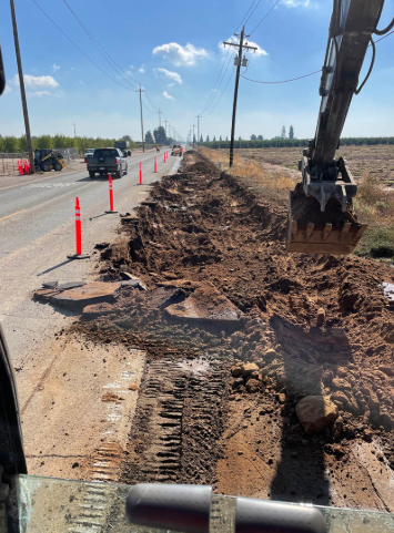 Roadwork trench beside a highway with an excavator digging and orange cones marking the lane