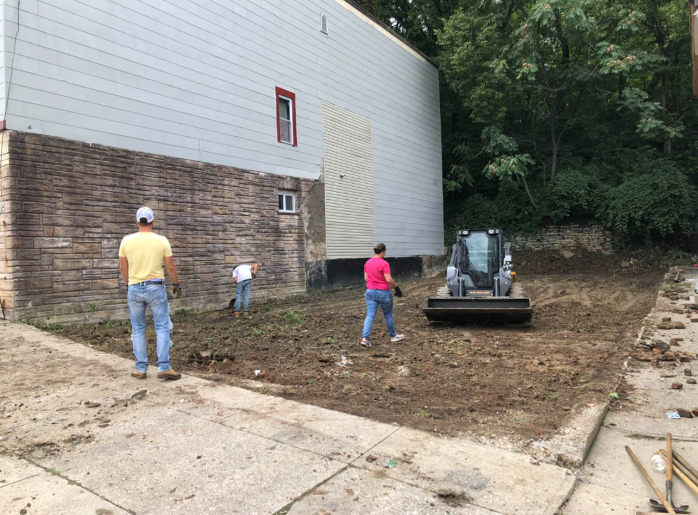Three people stand by a building with a muddy, cleared yard and heavy equipment nearby.
