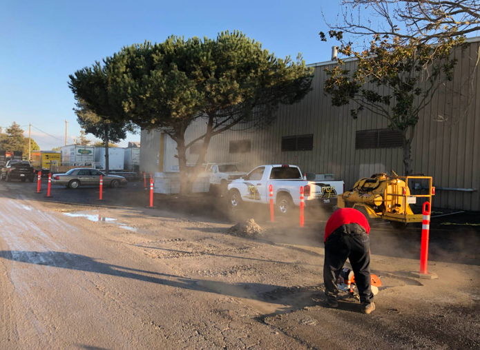Worker in red jacket repairing a muddy roadside near parked cars and orange cones