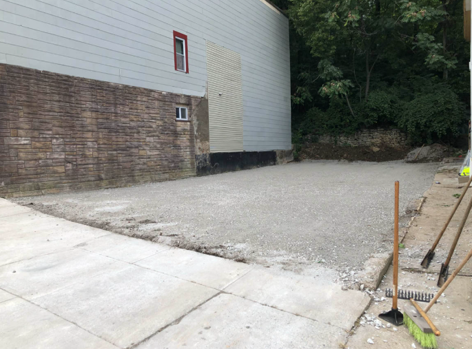 Empty gravel lot beside a brick and white building, bordered by trees and concrete paving.