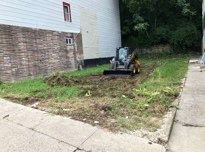 Small excavator clearing a muddy patch beside a building and sidewalk near trees
