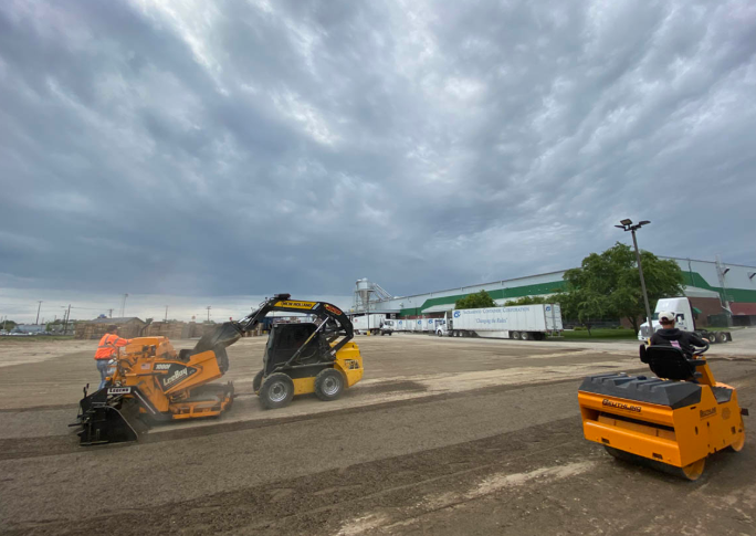 Orange skid-steers clearing debris on a road under a cloudy sky, with one worker on a machine at right.