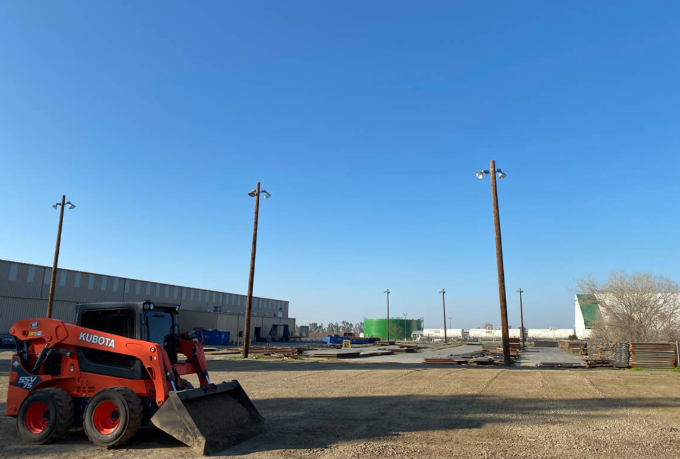 Orange skid steer loader on a dirt lot beside industrial buildings under a clear blue sky