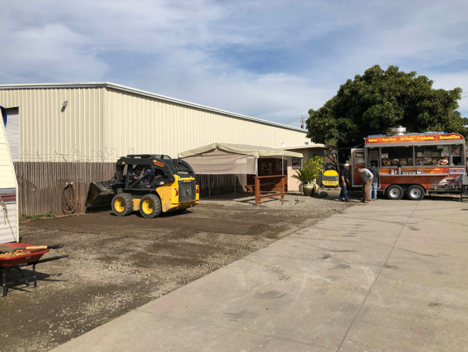 Construction yard with a skid steer, worker, and orange bus beside a warehouse