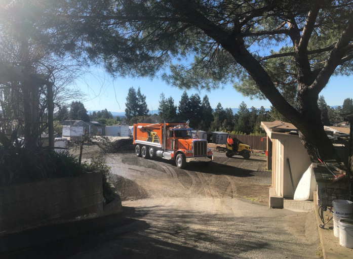 Orange dump truck and excavator on a dirt lot near trees and buildings