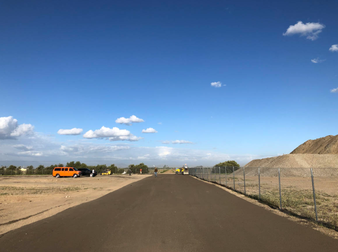 Paved road under a blue sky, with an orange truck, open field, and chain-link fence on the right.