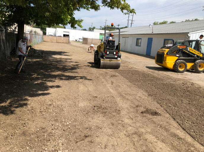 Construction yard with two skid steers on a dirt lot beside a long white building