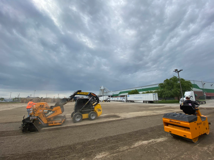 Construction site with three compact machines under a cloudy sky, including an orange loader and roller