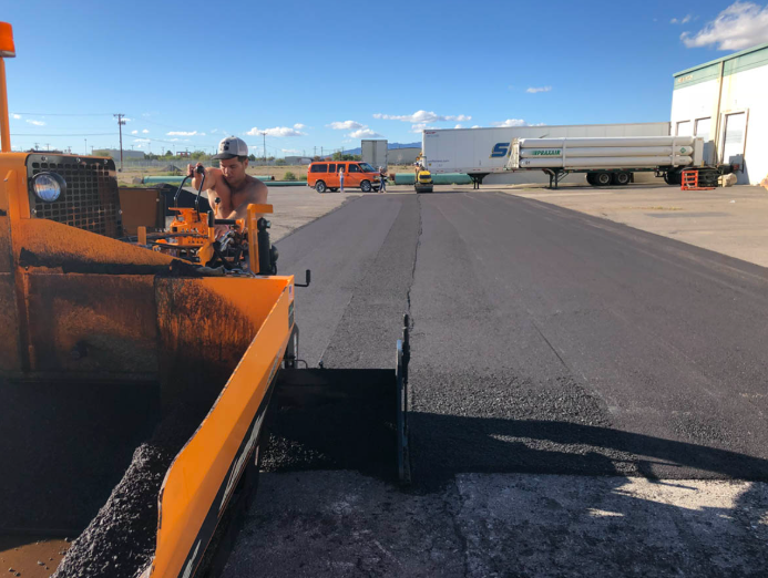 Worker operating an orange asphalt paver on a paved lot under a blue sky