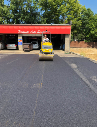 Small yellow road roller on fresh black pavement in front of a red-roofed garage with parked cars.