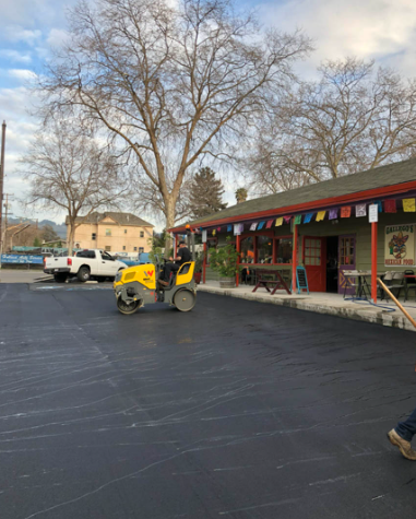 Yellow skid steer in parking lot beside red storefront with colorful flags and leafless trees.