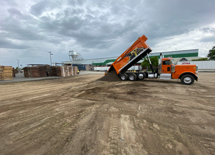 Orange dump truck unloading on a dirt lot under cloudy skies, with pallets stacked nearby.