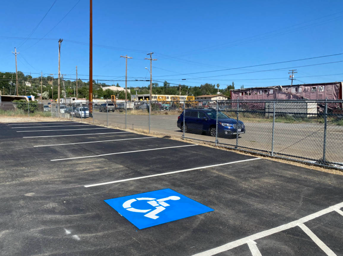 Accessible parking space in a mostly empty lot beside a chain-link fence and parked car.