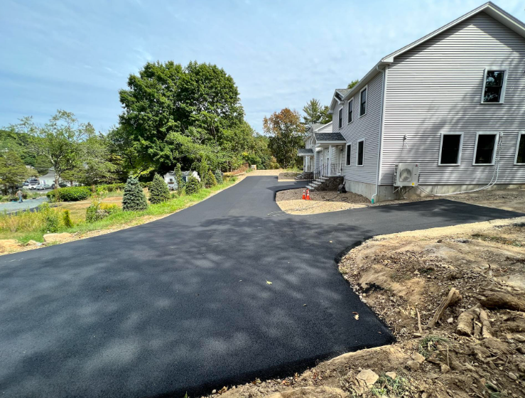 Freshly paved black driveway curving beside a light gray house and dirt yard under a blue sky