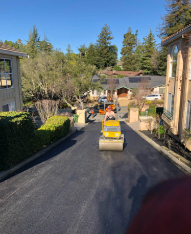 A small yellow ride-on roller on a newly paved driveway beside homes and trees.