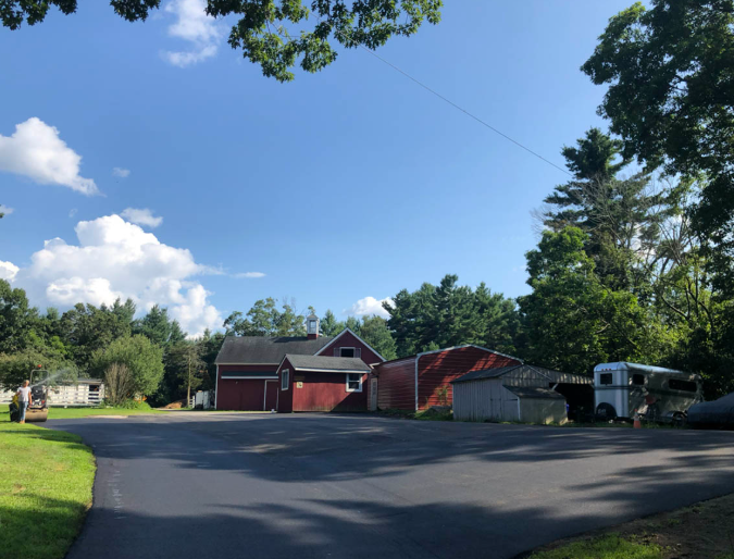 Driveway leading to red barns and outbuildings under a blue sky, framed by trees