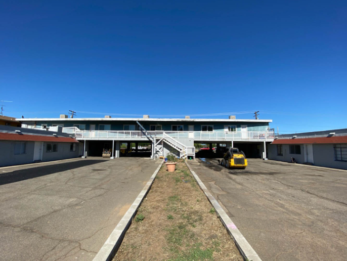 Motel-style building with central stairs, parking lot, and a yellow car under a clear blue sky