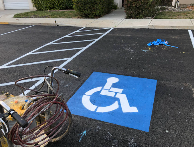 Damaged wheelchair parking sign painted on asphalt in a mostly empty parking lot, with cables and debris nearby