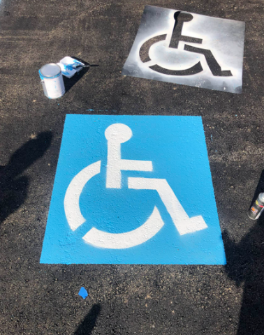 Two painted wheelchair accessibility symbols, one blue and one gray, on pavement near a paint can.