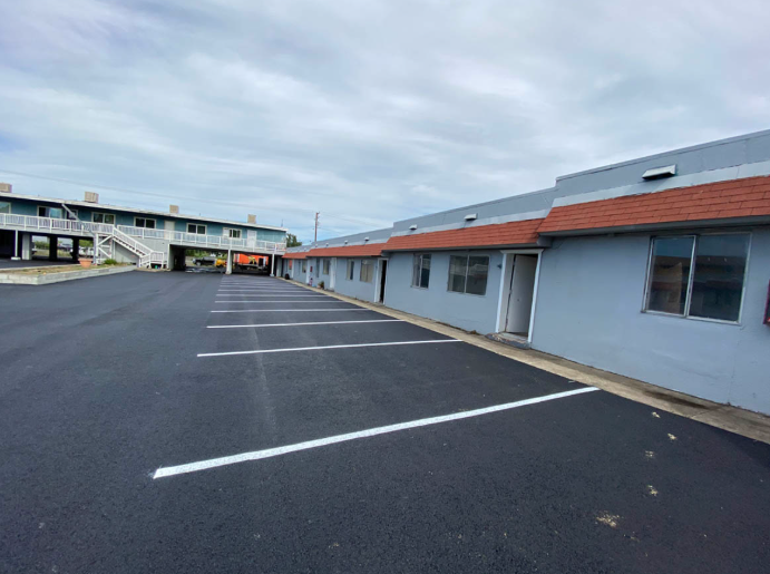 Empty motel parking lot beside a row of gray rooms under a cloudy sky