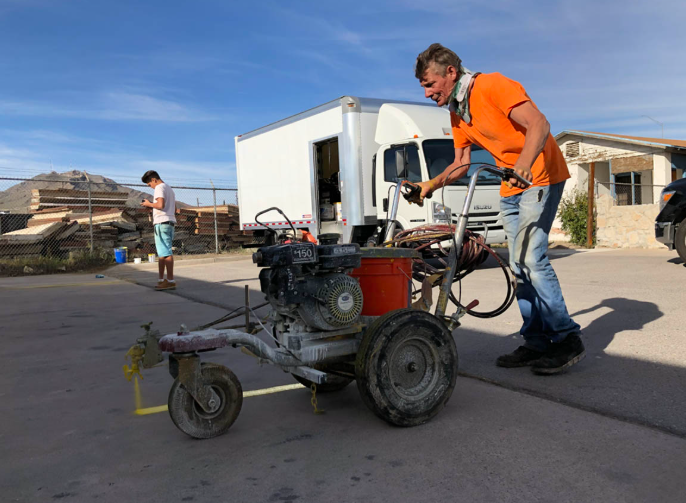 Man pushing a loaded cart outdoors in a lot with buildings and another person in the background
