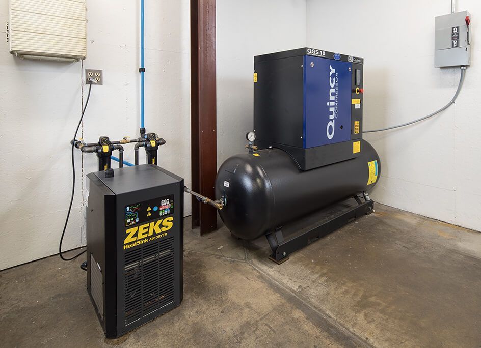 Industrial air compressor system with black tank and blue ZEKS unit in a utility room.