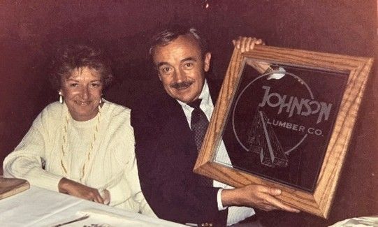A couple poses with a framed mirror featuring the logo for Johnson Lumber Co.
