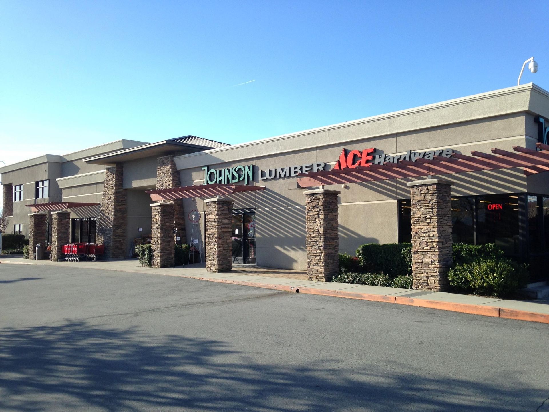 A tan, multi-unit building with stone pillars and a red, wooden pergola over the entrance for an Ace Hardware store.