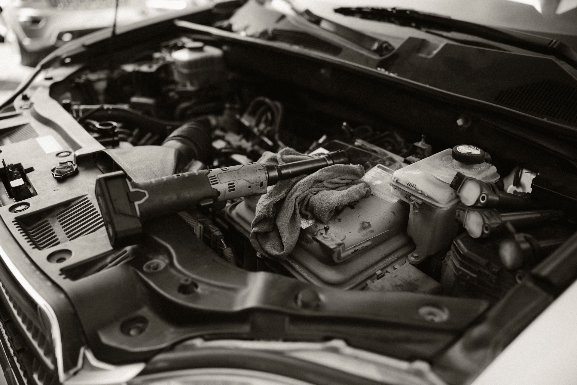 Black-and-white view of a car engine bay with the hood open and a mechanic’s gloved hand working inside