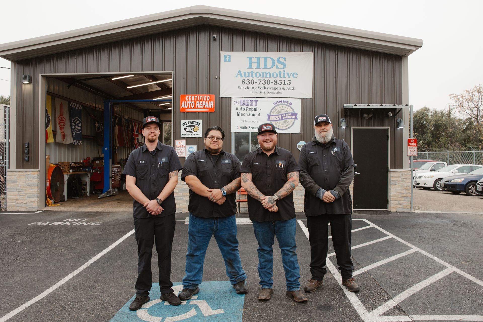 Four workers stand outside a warehouse garage with tools and vehicles visible inside.