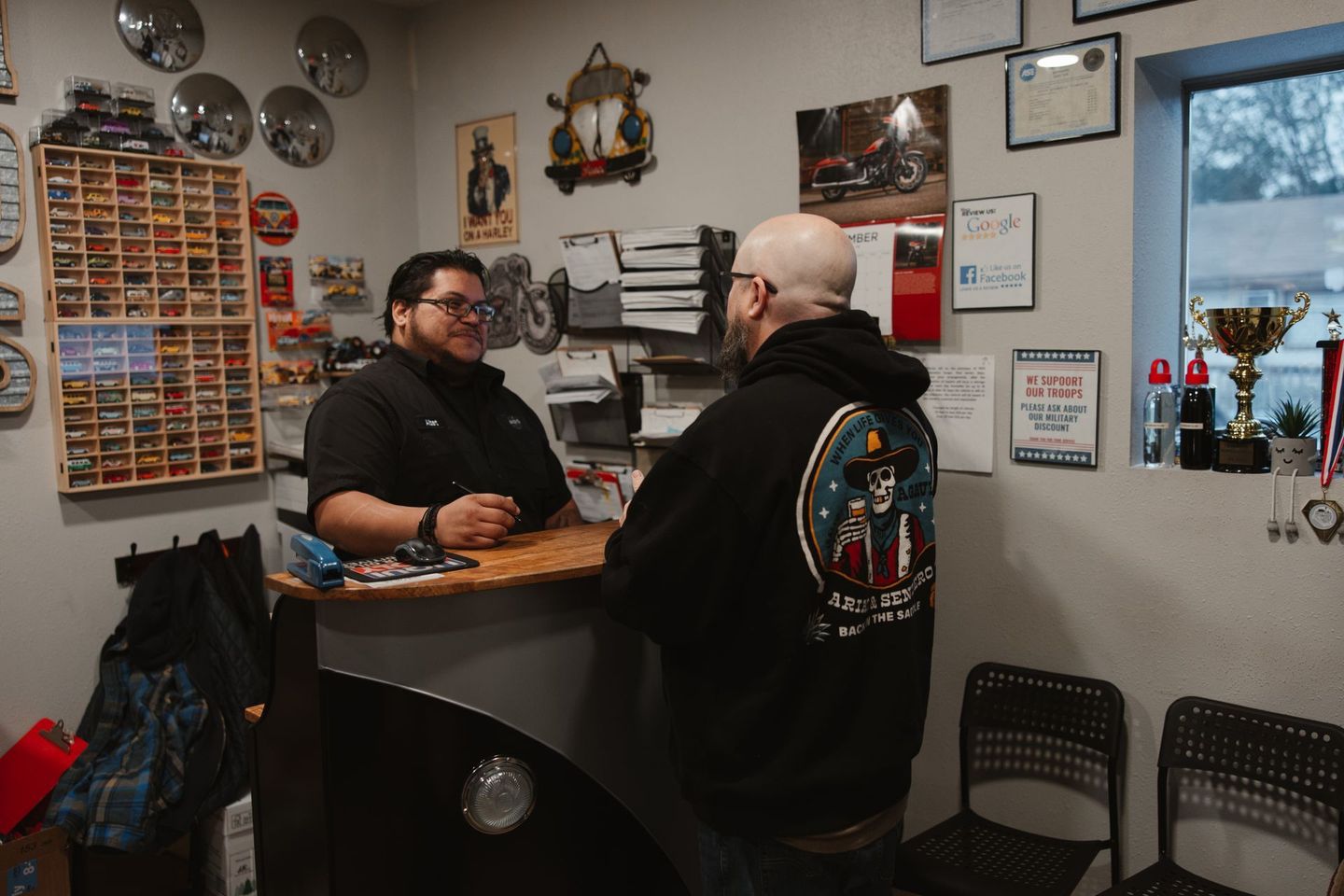 Two people talking across a counter in a small shop or café with wall decor and a window.