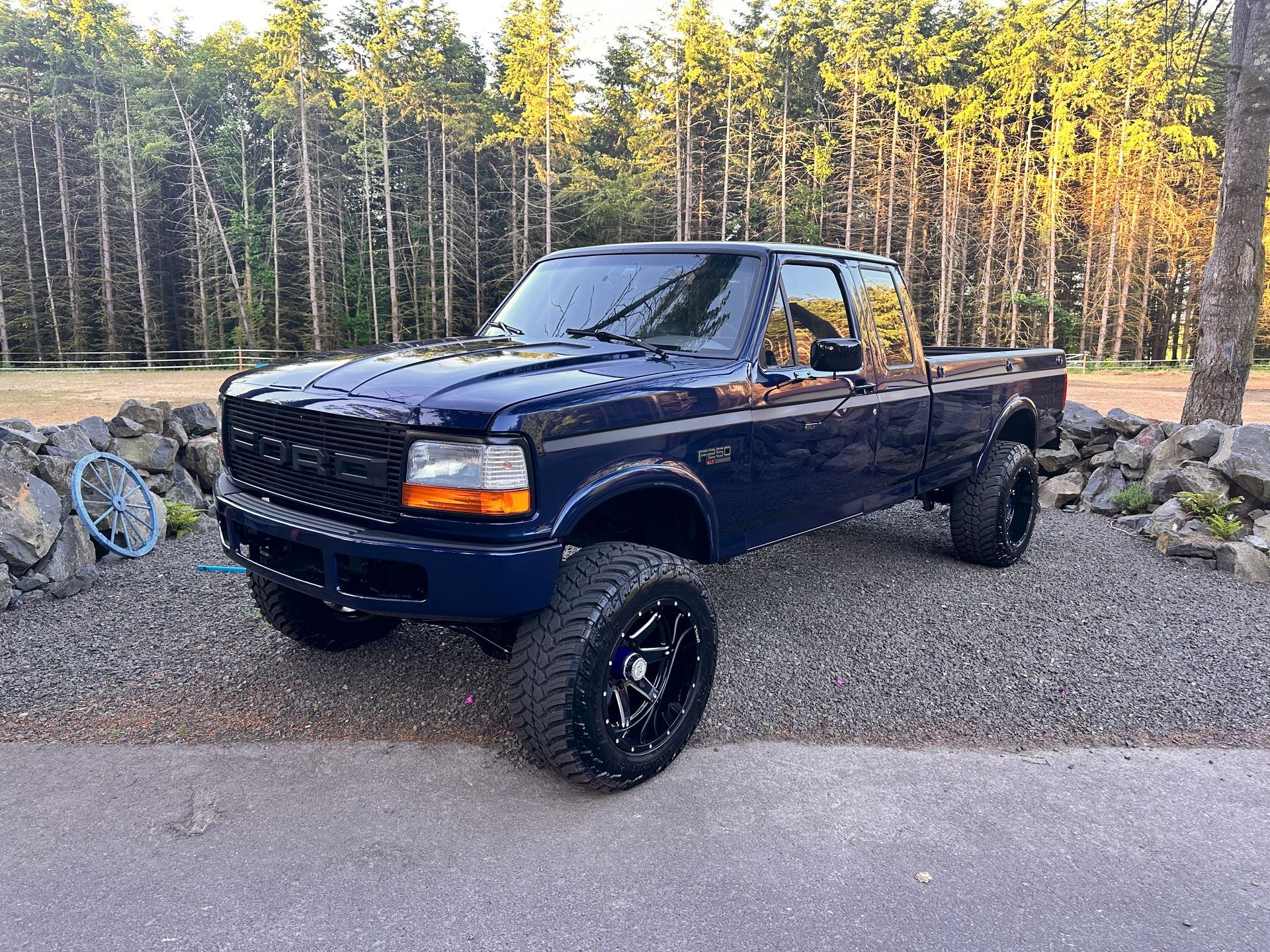 A blue truck is parked on a gravel road in front of a forest.