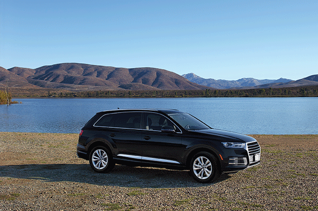 A black suv is parked on the shore of a lake with mountains in the background.