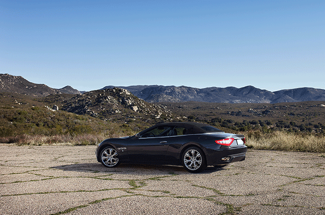 A black convertible sports car is parked in a dirt field with mountains in the background.