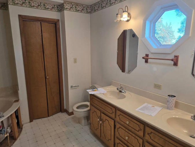 Bathroom with double vanity, octagonal window, toilet, and wood closet doors