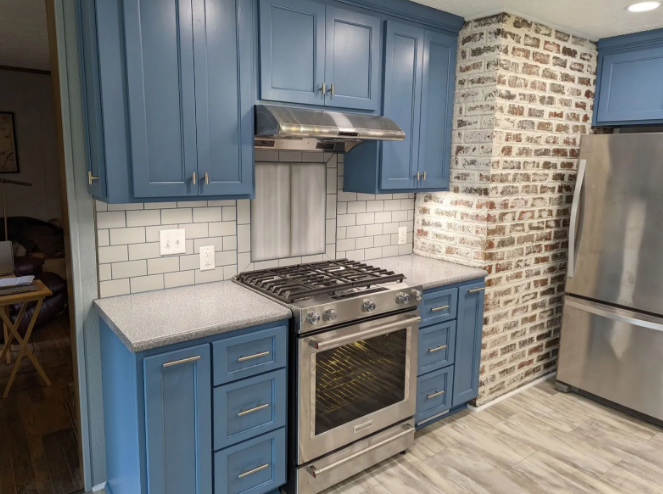 Blue kitchen cabinets with stainless steel stove and hood, white tile backsplash, and brick accent wall.