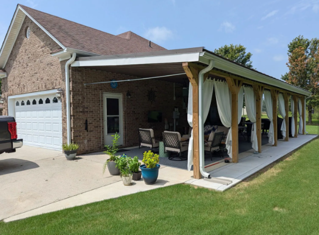 Brick house with attached white carport, patio seating, and potted plants beside a green lawn