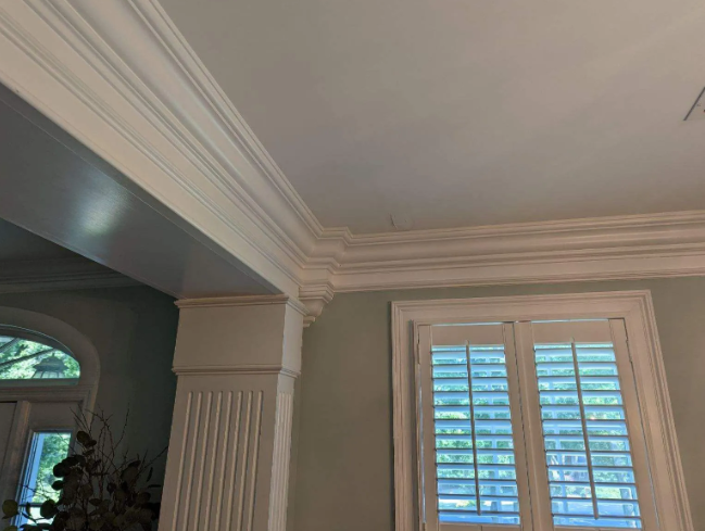Interior ceiling and crown molding above a window with white shutters in a gray room