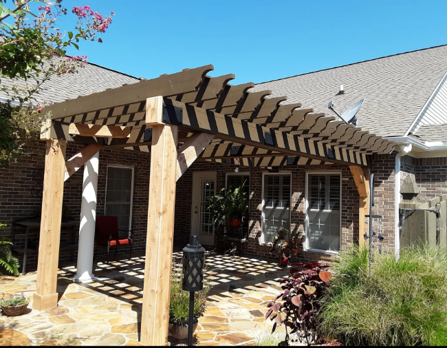 Wooden pergola over a stone patio beside a brick house, with potted plants and a pergola bench area