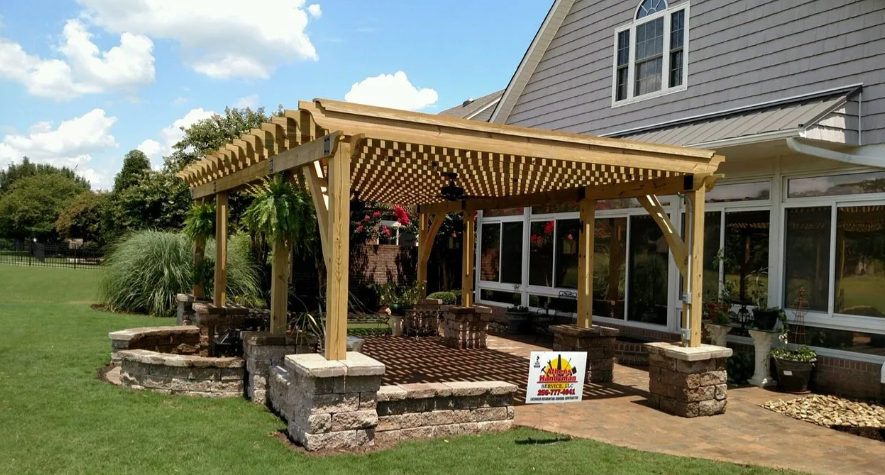 Wooden pergola over a landscaped patio beside a house, with stone pillars and flower beds.