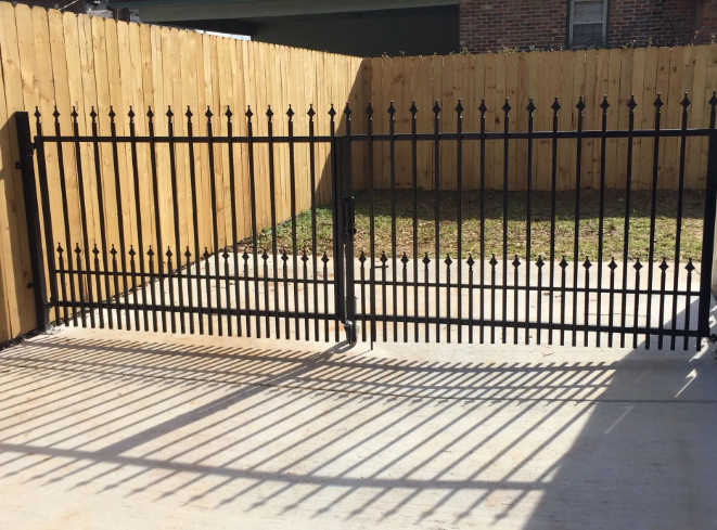 Black metal gate in front of a wooden fence, casting shadows on a concrete driveway.