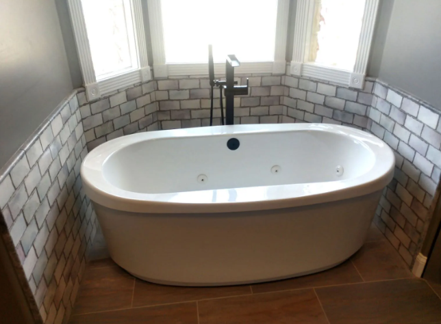 Freestanding white bathtub under three windows in a tiled bathroom